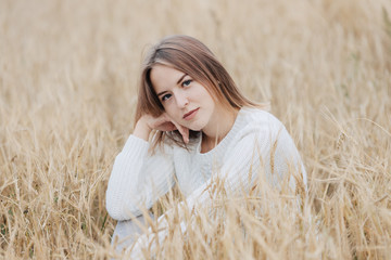 Beautiful young girl in a white sweater sits in a wheat field.