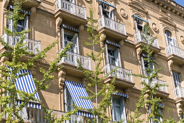 Fototapeta na wymiar Exterior of an elegant building with striped awnings on the balcony windows and young trees in front it 
