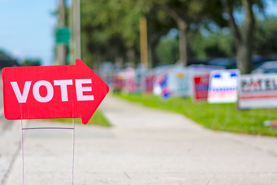 Sign directing voters to a polling place with a row of political signs in the background.