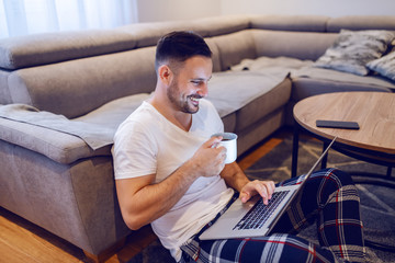 Beautiful caucasian unshaven smiling man sitting on floor in living room, surfing on internet and...