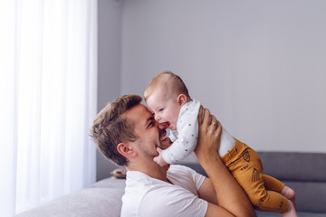 Playful handsome caucasian young dad lifting his loving 6 months old baby boy while sitting on sofa in living room. Baby enjoying time with father and laughing.