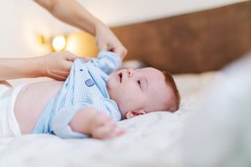 Close up of adorable baby boy lying on bed while his mother changing his clothes before bedtime. Bedroom interior.