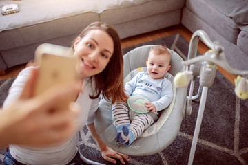 Top view of smiling proud caucasian mother taking selfie with her adorable baby boy. Baby sitting in rocker chair and smiling. Living room interior.