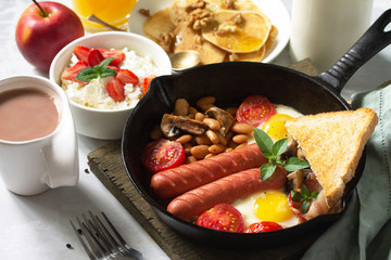 Keto breakfast: fried eggs, sausages, beans, bacon, mushrooms, grilled toasts, orange juice, cottage cheese, pancakes and cocoa on a light stone table.