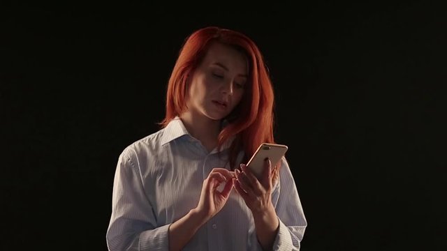 beautiful red-haired girl in a white shirt talking on a smartphone isolated on a black background. Phone advertising. Businesswoman in work