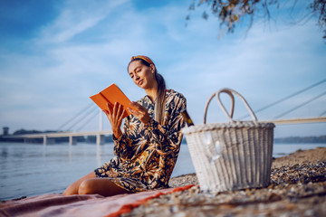 Attractive caucasian brunette in floral dress and with headband sitting on blanket on the shore and reading book. In foreground is picnic basket.