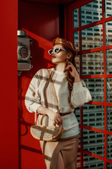 Outdoor autumn fashion portrait of young elegant woman wearing sunglasses, brown leather beret, white sweater, skirt, with beige small shoulder bag posing in red phone booth, call box