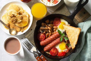 Keto breakfast: fried eggs, sausages, beans, bacon, mushrooms, grilled toasts, orange juice, cottage cheese, pancakes and cocoa on a light stone table. Top view flat lay background.