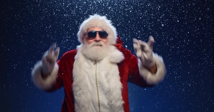 Cool santa claus wearing sunglasses dancing to music, isolated over snowy blue background - christmas party, christmas spirit concept close up 4k footage