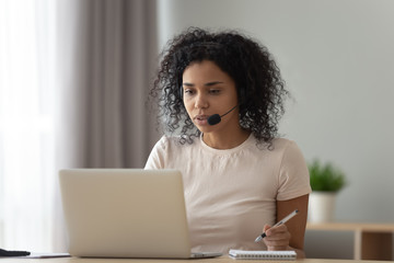 Focused African American woman in headset using laptop, writing notes