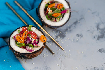 Fresh vegetables salad in coconut peel. Tomatoes, spinach, peppers, chili, radishes. On a gray, stone background. Chopsticks. Copy space. Vegetarian, healthy food. 