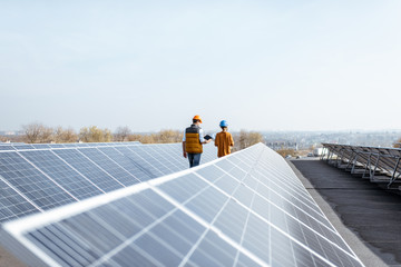 View on the rooftop solar power plant with two engineers walking and examining photovoltaic panels....