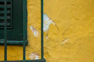 A wooden green window with a green fence in front and a yellow wall with some peeling white parts appearing
