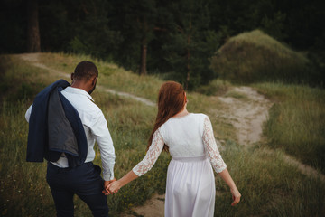 Naklejka na ściany i meble Back view of interracial going couple. walking friendly girl and guy holding hands. Rear view people collection. Multi-ethnic couple holding hands goes