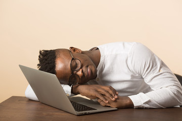 handsome young african man in glasses and a white shirt sits at a table with a laptop on a beige background