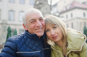 Handsome elderly man is embracing his young blonde wife spending time together outdoors in the ancient city during early spring or autumn.