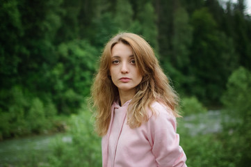 Serious young blonde woman in pink clothes with long hair is looking at the camera with anxiety on the hill with green forest background during spring or early autumn in the mountains.