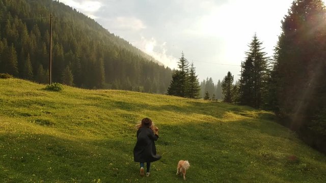 cinema shot of aerial photography hipster girl in coat and hat with her dog red border collie runs on a green field in a canyon among the mountains