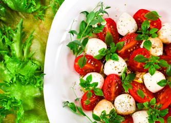 Caprese salad. Ripe red cherry tomatoes, mozzarella cheese balls, young basil leaves and spices with leaves of frills lettuce. Top view.