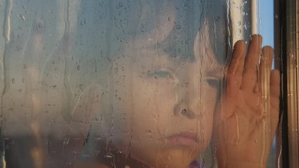 Child loneliness in the rain. Sad little girl looks out the rainy window. Quarantine concept.