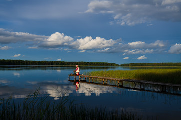 View of a large lake surrounded by forest on a sunny summer day. A woman fisherman sits on a wooden platform. Blue sky and clouds are reflected in the water. Scenery. Nature.