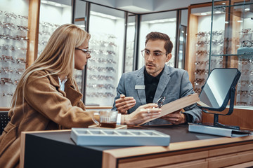 Woman is choosing eyeglasses while male optician sitting near with another eyeglasses.