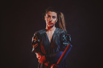 Beautiful woman is posing for photographer with baseball bat at dark photo studio.