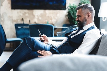 Smiling bearded man making text message on smartphone while sitting sofa at modern office