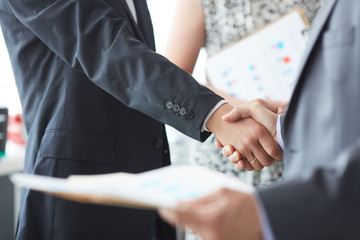 Business people in suits shaking hands, finishing up a meeting. Male hands in handshake closeup with male colleague in background.