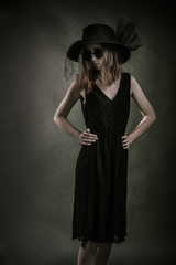 Studio portraitgirl black dress, hat and sunglasses stands on a dark background