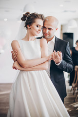 Gorgeous bride and groom gently hugging and dancing at wedding reception in restaurant. Beautiful stylish wedding couple performing first dance and embracing. Sensual romantic moment