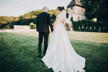 Gorgeous bride and groom walking in sunset light. Beautiful stylish wedding couple embracing in summer park, back view. Sensual romantic moment. Newlyweds