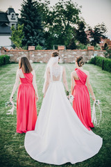 Fototapeta premium Gorgeous bride walking with bridesmaids in pink dresses, holding stylish bouquets in warm evening light in summer park. Beautiful happy bride with maid of honor posing together