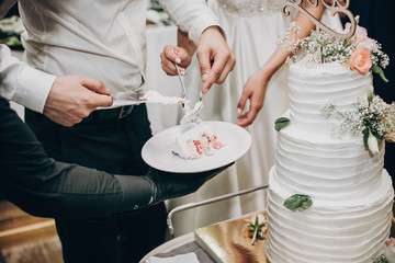 Obraz na płótnie Canvas Bride and groom cutting stylish wedding cake at wedding reception in restaurant. Wedding couple holding slice of wedding cake decorated with flowers