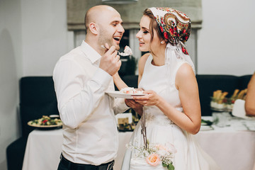 Bride and groom tasting stylish wedding cake at wedding reception in restaurant. Wedding couple plate with slice of cream cake decorated with flowers and eating