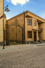 Wrought iron gate with monogram near a modern building of light tuff with wrought iron balcony in a...