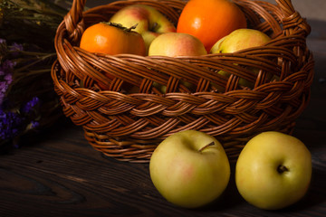 a wicker basket filled with fruits, persimmons and apples, in front of the basket lies two apples. near lie wildflowers 