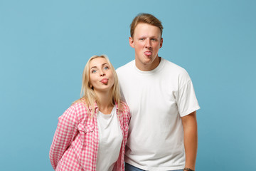 Fototapeta premium Young cheerful couple two friends guy girl in white pink empty blank design t-shirts posing isolated on pastel blue background in studio. People lifestyle concept. Mock up copy space. Showing tongue.