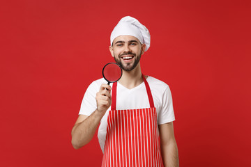 Smiling young bearded male chef cook or baker man in striped apron white t-shirt toque chefs hat isolated on red wall background. Cooking food concept. Mock up copy space. Holding magnifying glass.