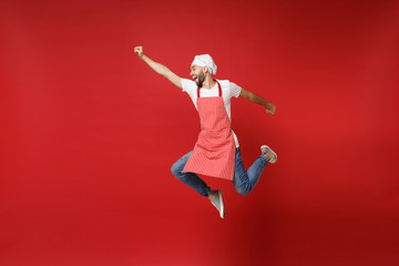 Funny young male chef cook or baker man in striped apron white t-shirt toque chefs hat posing isolated on red background. Cooking food concept. Mock up copy space. Jumping, fooling around, having fun.