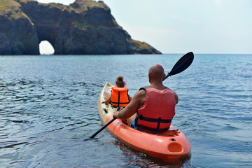 A father and his little daughter are sailing on the sea in a kayak to a grotto in the rock.