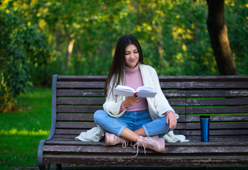 Obraz premium young female is reading a book on the bench in the park, thermo mug on the bench