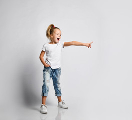 Happy screaming kid girl in blue jeans, white t-shirt and sneakers is looking at something and pointing at it on white