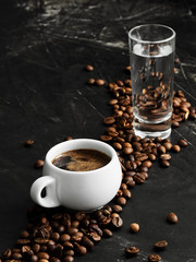 White cup with a strong espresso on a black background. Fragrant freshly brewed coffee. Roasted coffee beans are located around the cup. Next to a cup of coffee is a glass of cold water. Close-up.