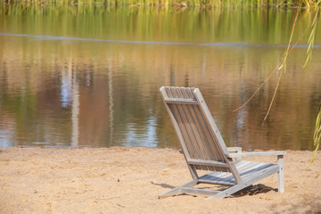 Dreamy scene of wooden beach chair in sand with reflections of reeds in water and willow branches on side - selective focus.