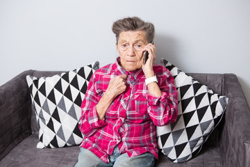An old elderly woman grandmother with gray hair sits at home on the couch using the hand phone, a...
