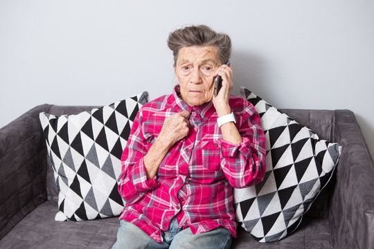 An Old Elderly Woman Grandmother With Gray Hair Sits At Home On The Couch Using The Hand Phone, A Telephone Conversation To Hear The Bad News. Emotion Fear Scare