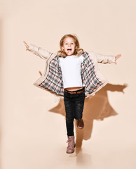 Joyful blonde kid girl in opened beige raincoat is running jumping with her hands spread like wings, happy screaming