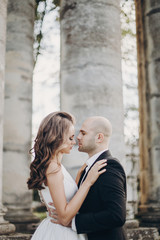 Gorgeous bride and groom embracing in evening sunlight near old castle in park. Romantic moment. Stylish wedding couple gently hugging and smiling