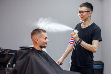 young Kazakh barber works in a barbershop, a young guy makes a short haircut at a hairdresser, wet his head, water splashes on a gray wall background